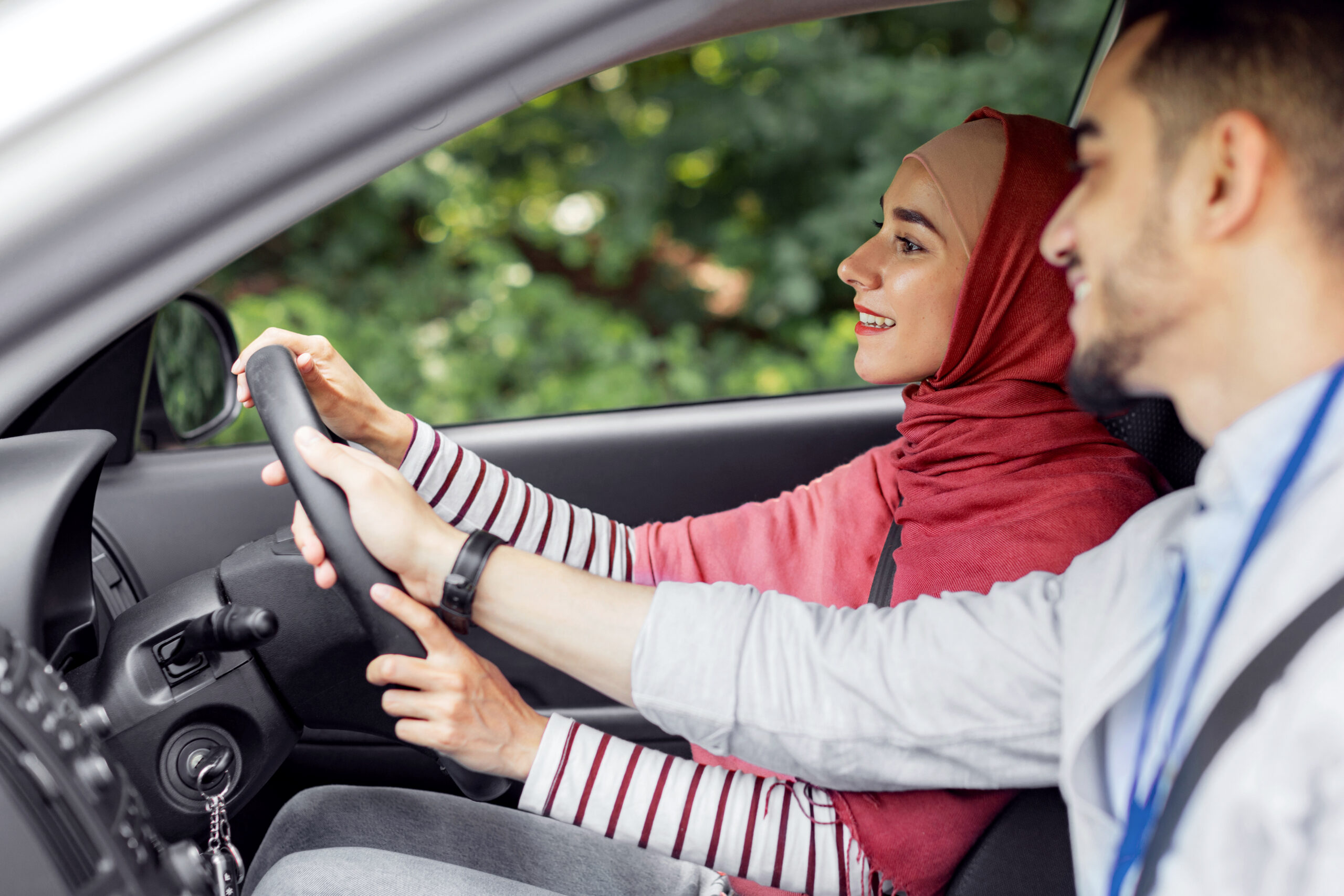 Happy attractive millennial arabic man teaching student, helping to woman in hijab at steering wheel to driving car