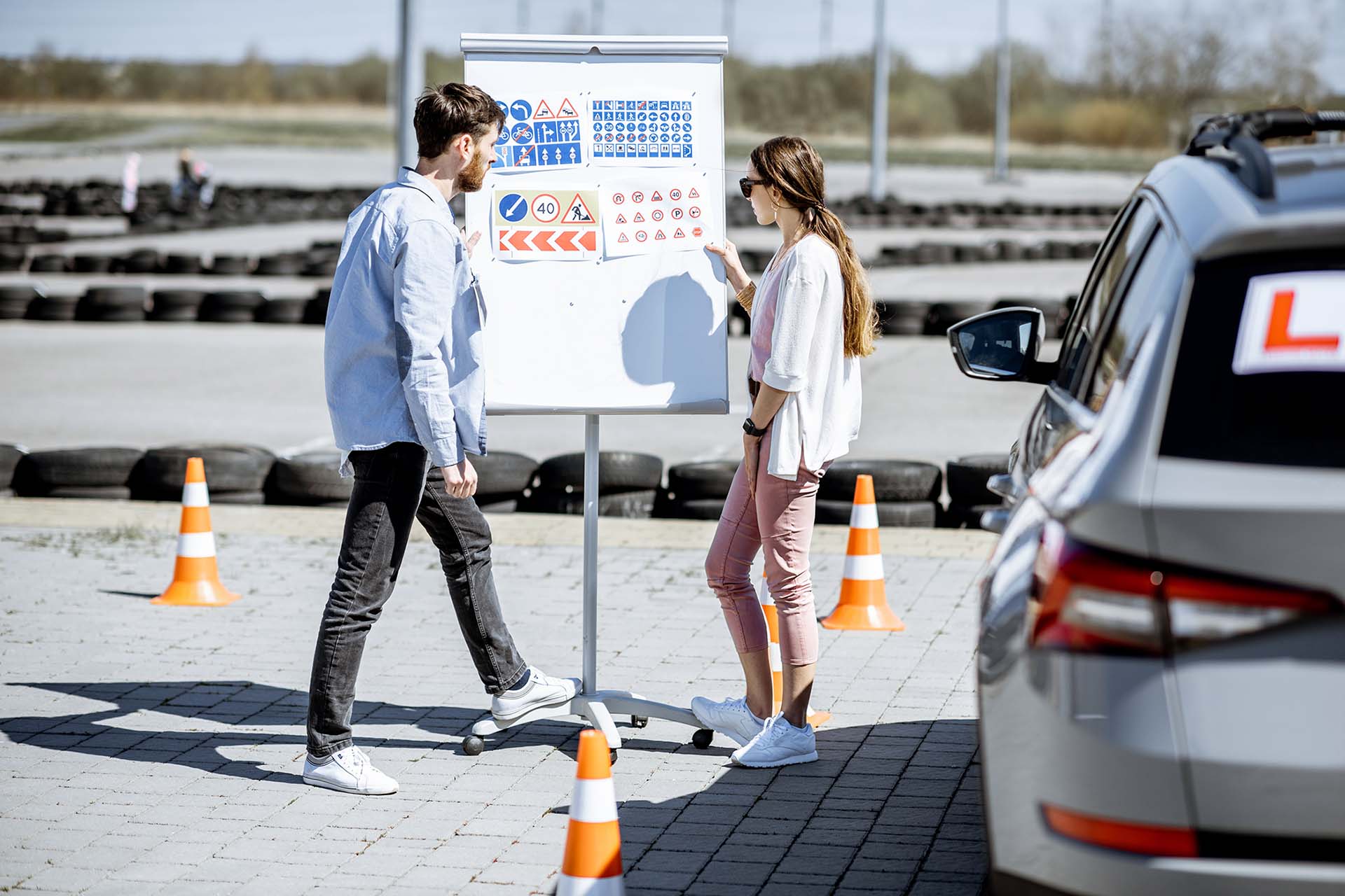 Two people standing beside a presentation board covered with road signs in a driving practice area, discussing the signs near a learner‑driver car and traffic cones.