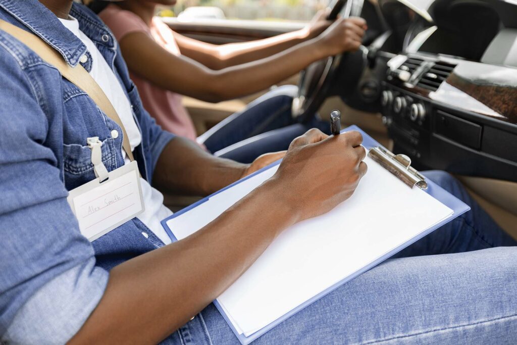 Person in the passenger seat holding a clipboard and pen with a visible name tag, taking notes while a student driver holds the steering wheel during a driving test inside a car.